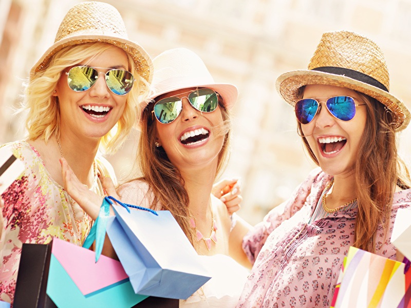 Drie vrolijke vrouwen met zonnebrillen en strohoeden genieten van een dagje shoppen, met kleurrijke tassen in hun handen.