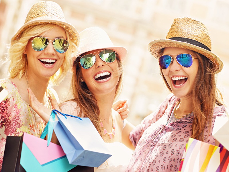 Three cheerful women with sunglasses and straw hats are enjoying a day of shopping, with colorful bags in their hands.