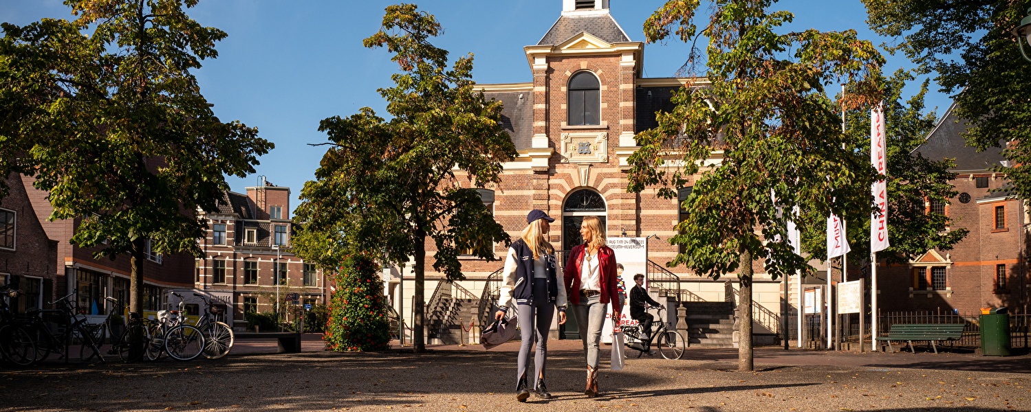 Twee vrouwen lopen met shoppingbags over een zonnig plein in Hilversum, met op de achtergrond het Museum Hilversum en banners van een expositie.