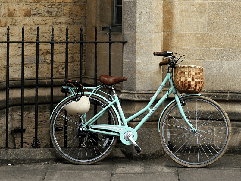 Lichtgroene damesfiets met een rieten mand, geparkeerd tegen een stenen muur naast een zwart hek.