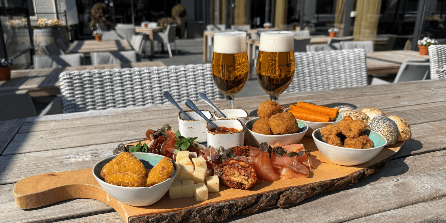 Plateau Amrâth geserveerd op een houten plank met diverse luxe hapjes zoals kaasblokjes, charcuterie, bitterballen, kipstukjes, dips en broodjes, vergezeld van twee glazen bier op een zonnig terras.