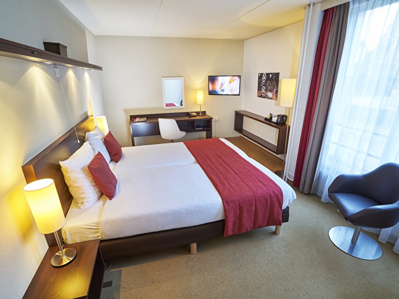 Comfort kamer in Amrâth Hotel Media Park Hilversum met een tweepersoonsbed met rode kussens, rood sprei een zwarte stoel en een televisie aan de muur