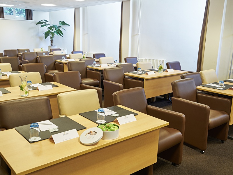 Vergaderruimte met moderne opstelling, voorzien van comfortabele stoelen, houten tafels met notitieblokken, waterglazen en een schaal met groene appels.
