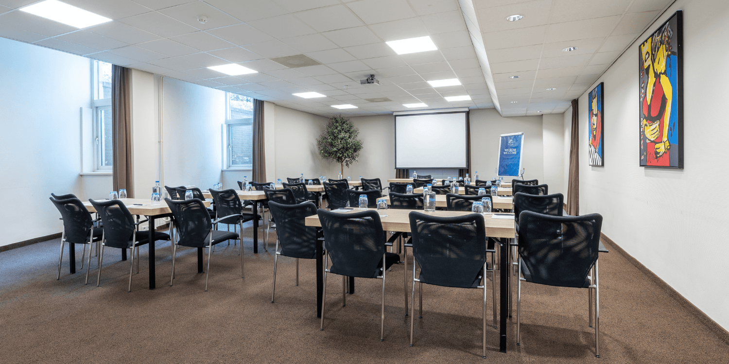 Spacious meeting room with comfortable brown and beige chairs, wooden tables equipped with notebooks, pens, water glasses and decanters and with decorative plants and colorful art on the wall. 